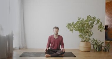 Kafkasyalı bir adam, güneşli bir dairede bağdaş kurarak meditasyon yapıyor. Sakallı yoga eğitmeninin portresi sukhasana asana yavaş çekimde dinleniyor. Rahatlama stresi üstesinden gelir.