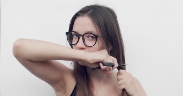 Femme avec des lunettes coupe ses cheveux sur fond blanc. Jeune femme coupe ses longs cheveux bruns avec des ciseaux par elle-même pendant l'auto-isolement. Changement de coiffure pour surmonter le stress et le concept de soins capillaires — Video