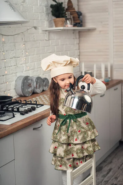 little girl in a chef's cap examines a kettle in the kitchen