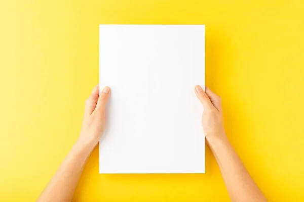 Young woman hands showing blank paper sheet on yellow desktop. Top view