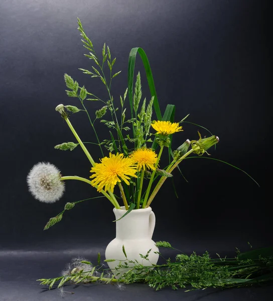 Küçük seramik sürahi ve karahindiba tohum sarı, yarı çiçek açması ve kabarık dandelions ve çeşitli çayır çimen ve siyah arka plan üzerine biraz ot bir yüzey üzerinde bulunan