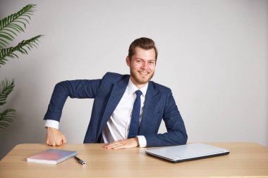 Mavi takım elbiseli bir işadamı masasında oturmuş elini yumruğuyla tutuyor. Doğrudan kameraya bakıyor ve gülümsüyor. Beyaz arkaplan.