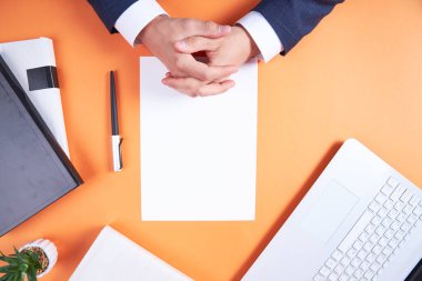 İş aksesuarları olan turuncu bir masaüstü, yukarıdan görüş. Beyaz bir kağıt, bir kalem, bir kitap. İş adamı kollarını kavuşturdu. Ticari aksesuarları olan düz bir ev..
