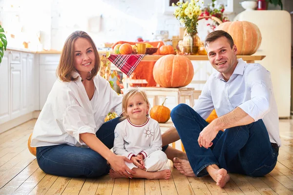 A happy family of three, mother, father and daughter, stand on a light kitchen, a wooden table with an autumn harvest - pumpkins, apples — Stock Photo, Image