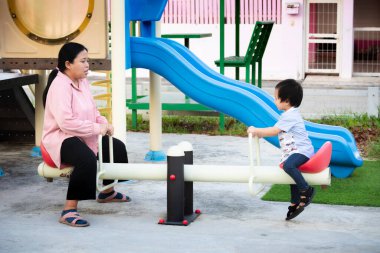 Küçük mutlu çocuk, annesiyle parkta tahterevalli oynarken eğleniyor. Açık hava parkında bir anne ve oğlu arasında eğlenceli bir an, oyun zamanının tadını çıkarıyor. 2-3 yaşlarında bir çocuk..