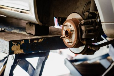 Eski, yıpranmış bir fren diski ve bir arabanın kalibreli görüntüsü. Araç mavi bir hidrolik asansörle kaldırılır, garajın içinde, araba tamiri ya da bakım ihtiyacını vurgular..