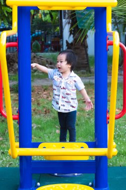 Küçük Asyalı çocuğun samimiyeti renkli mavi ve sarı bir ekipman makinesinin arkasında heyecanlı bir şekilde duruyor. Çocukluk oyununda neşeli ve aktif bir an. Çocuk açık havada yeni şeyler keşfediyor..