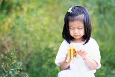 Şirin küçük kız soyulmuş bir muz tutuyor ve yemeye hazırlanıyor. Açık havada, çocuğun sağlıklı atıştırmanın tadını çıkardığı sessiz bir an. Yeşil çimenlerin bulanık arkaplan alanı, doğal ve sağlıklı renklerde..