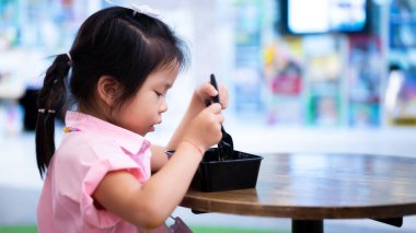 Saçları örgülü, yuvarlak ahşap bir masada oturan ve siyah bir kaşıkla yemek yiyen Asyalı bir çocuğun yan görüntüsü. Kız yemeklerine odaklanmış. Geçmişi canlı bir şekilde bulanıklaşmış..
