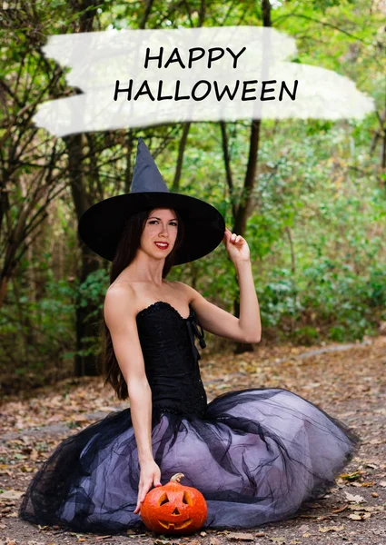 Halloween holiday background. Halloween Witch with a halloween pumpkin jack o lantern decor with funny face in a dark forest. Beautiful young woman in witches hat and costume holding pumpkin with text Happy Halloween