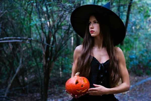 ハロウィーンの休日背景 暗い森で変な顔とハロウィーン カボチャのジャック ランタン装飾が施されたハロウィーンの魔女 魔女帽子とカボチャを保持している衣装の美しい若い女性 — ストック写真