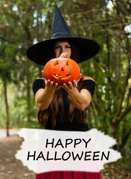 Halloween holiday background. Halloween Witch with a halloween pumpkin jack o lantern decor with funny face in a dark forest. Beautiful young woman in witches hat and costume holding pumpkin with text Happy Halloween