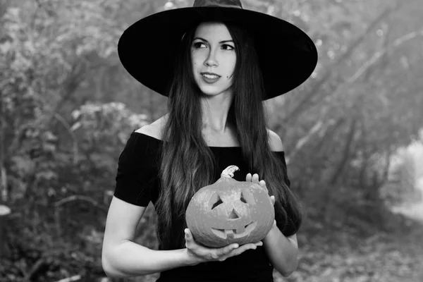 Halloween holiday background. Halloween Witch with a halloween pumpkin jack o lantern decor with funny face in a dark forest. Beautiful young woman in witches hat and costume holding pumpkin. Black and white photo.