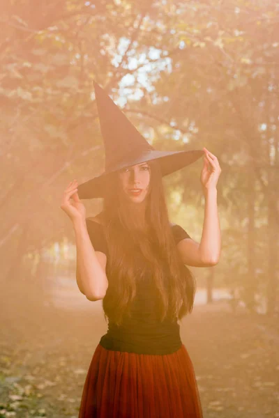 ハロウィーンの休日背景 暗い森のハロウィン魔女 魔女帽子とオレンジ色の煙背景に衣装の美しい若い女性 — ストック写真