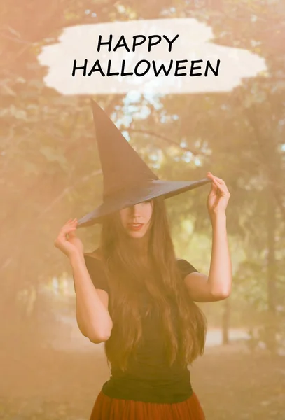 Halloween holiday background. Halloween Witch in a dark forest. Beautiful young woman in witches hat and costume on orange smoke background with text happy halloween.