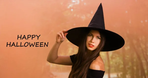 Halloween holiday background. Halloween Witch in a dark forest. Beautiful young woman in witches hat and costume on orange smoke background with text happy halloween.