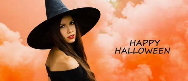 Halloween holiday background. Halloween Witch in a dark forest. Beautiful young woman in witches hat and costume on orange smoke background with text happy halloween.