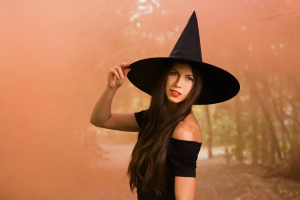 ハロウィーンの休日背景 暗い森のハロウィン魔女 魔女帽子とオレンジ色の煙背景に衣装の美しい若い女性 — ストック写真