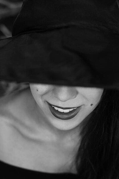 Halloween holiday background. Halloween Witch in a dark forest. Close-up face with red lips. Beautiful young woman in witches hat and costume. Black and white photo.