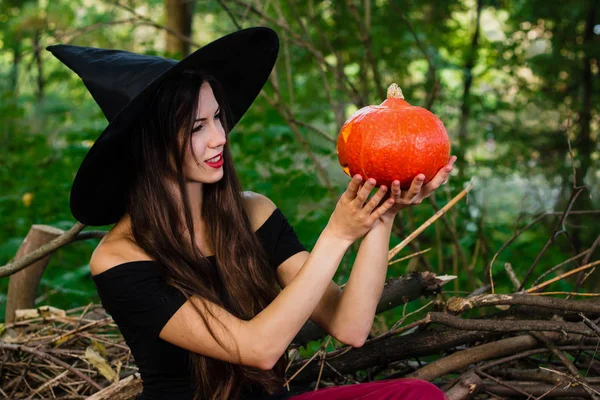 Halloween holiday background. Halloween Witch with a halloween pumpkin jack o lantern decor with funny face in a dark forest. Beautiful young woman in witches hat and costume holding pumpkin.