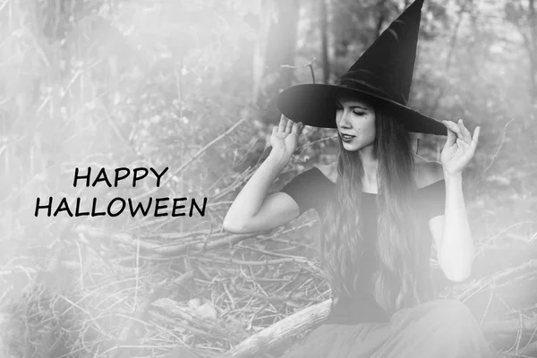 Halloween holiday background. Halloween Witch in a dark forest with toned effect. Beautiful young woman in witches hat and costume with text happy halloween. Black and white photo.