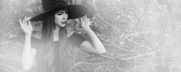 Halloween holiday background. Halloween Witch in a dark forest with toned effect. Beautiful young woman in witches hat and costume. Black and white photo.