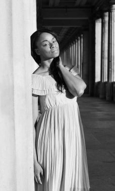 Genç güzel kadın olumlu kameraya gülümseyen afro kıvırcık siyah saçlı siyah. Nazik bej elbise ve güneşin doğuşunu büyük holü duran bir kadın. Siyah ve beyaz fotoğraf.