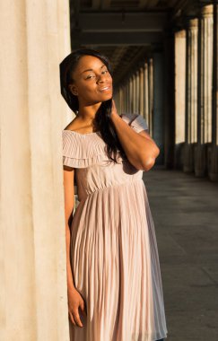 Genç güzel kadın olumlu kameraya gülümseyen afro kıvırcık siyah saçlı siyah. Nazik bej elbise ve güneşin doğuşunu büyük holü duran kadın. 