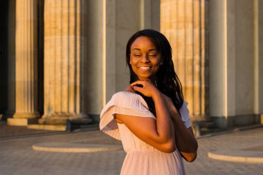 Genç güzel kadın olumlu ile kapalı gözler gülümseyen afro kıvırcık siyah saçlı siyah. Nazik bej elbise ve güneşin doğuşunu büyük holü duran kadın. 