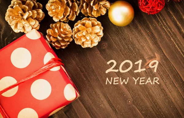 Christmas and New Year\'s Day festive decoration, golden ball, golden fir cones with present wrapped in red paper with golden circles on brown wood background. Flat lay. View from above with text 2019 new year.