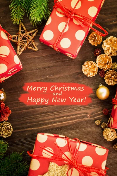 Christmas and New Year's Day festive decoration, red and golden balls, fir cones, branches and wooden stars with presents wrapped in red paper with golden circles on brown wood background with text Merry Christmas and Happy New Year on red. Flat lay.