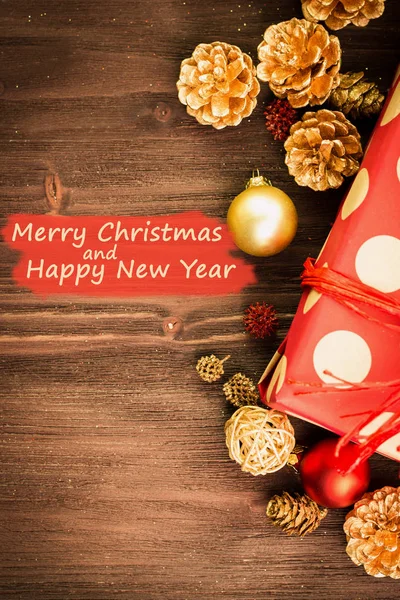 Christmas and New Year\'s Day festive decoration, golden ball, golden fir cones with present wrapped in red paper with golden circles on brown wood background. Flat lay. View from above with text Merry Christmas and Happy New Year on red.