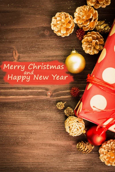 Christmas and New Year's Day festive decoration, golden ball, golden fir cones with present wrapped in red paper with golden circles on brown wood background. Flat lay. View from above with text Merry Christmas and Happy New Year on red.