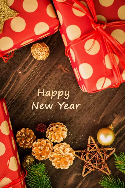 Christmas and New Year\'s Day festive decoration, golden balls, multicolored fir cones and branches with presents wrapped in red paper with golden circles on brown wood background. Flat lay. View from above with text happy new year.