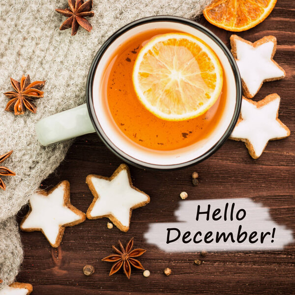 Winter theme. Hot tea with spices, orange, anise, cookies in a shape of star, pepper and gray scarf on wooden background. Flat lay, View from above with text hello December.