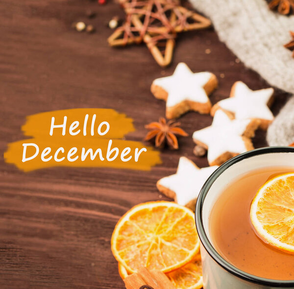 Winter theme. Cup of hot tea with spices, orange, cinnamon, anise, cookies in a shape of star, pepper and gray scarf on wooden background with text hello december. View from above.