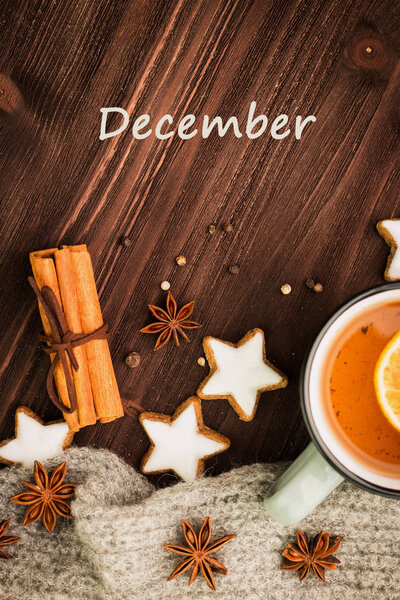 Winter theme. Hot tea with spices, orange, cinnamon, anise, cookies in a shape of star, pepper and gray scarf on wooden background. Flat lay, View from above with text december.