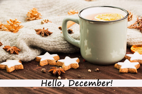 Winter theme. Cup of hot tea with spices, orange, cinnamon, anise, cookies in a shape of star, fir cones, pepper and gray scarf on wooden background with text hello december.