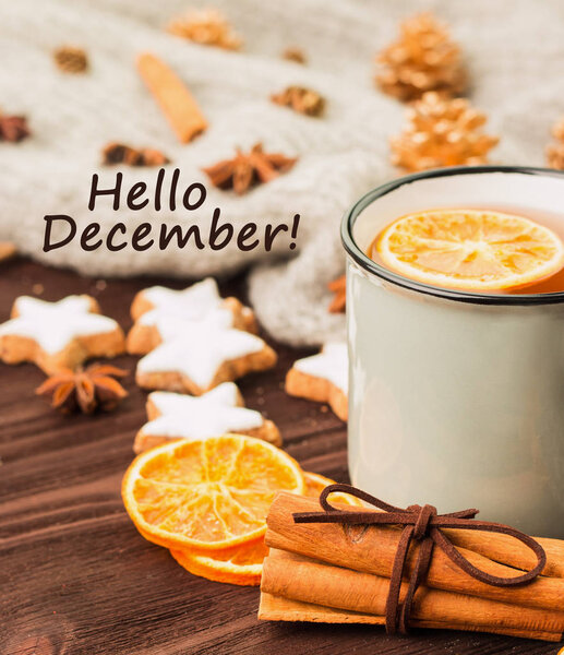 Winter theme. Cup of hot tea with spices, orange, cinnamon, anise, cookies in a shape of star, fir cones, pepper and gray scarf on wooden background with text hello december.