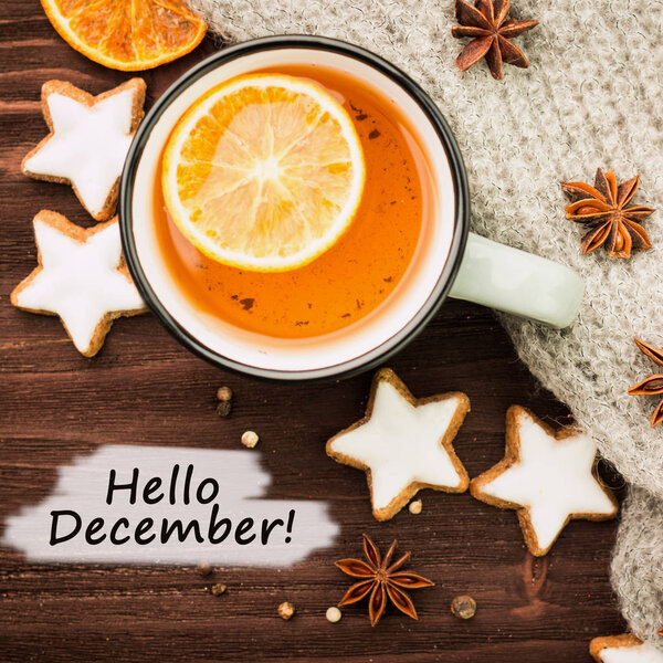 Winter theme. Hot tea with spices, orange, anise, cookies in a shape of star, pepper and gray scarf on wooden background. Flat lay, View from above with text hello December.