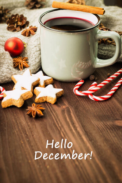 Winter theme. Hot steaming cup of glint wine with spices, cinnamon, anise, cookies in a shape of star, red candies, pepper and gray scarf on wooden background with text Hello December.