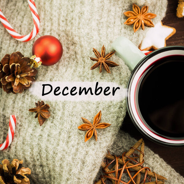 Christmas hot steaming cup of glint wine with spices, cinnamon, anise, cookies in a shape of star, red candies, fir cones, pepper and gray scarf on wooden background with text December. Flat lay. View from above.