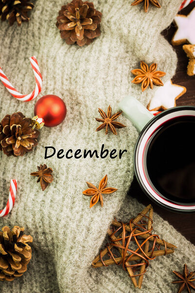 Christmas hot steaming cup of glint wine with spices, cinnamon, anise, cookies in a shape of star, red candies, fir cones, pepper and gray scarf on wooden background with text December. Flat lay. View from above.