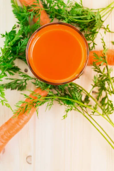 Fresh carrot juice in glass with carrots and green carrot tops on wooden table background. Healthy lifestyle concept. View from above. Flat lay. Copy space for text.