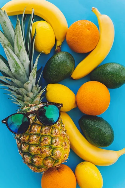 stylish pineapple in sunglasses and bananas,oranges,lemons,avocado on blue trendy paper background, flat lay. summer multi fruit concept, healthy lifestyle and party vibes