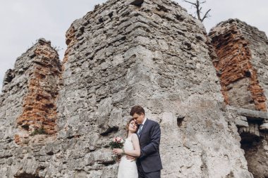 Eski kale duvarının yanında poz veren yeni evli çift, antik kale açıklarında peri masalı düğünü, gelin ve damadın kale kulesinin yanında kucaklaşması, düğün anı.