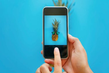 instagram meyve fotoğraf. telefon ve ananas alarak fotoğraf güneş gözlüğü mavi kağıt üzerinde tutan el, trendy düz koymak. şık gıda Fotoğrafçılık. yaratıcı telefon görüntü
