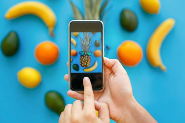 El telefon ve ananas güneş gözlüğü ve muz, portakal ve avokado alarak fotoğraf mavi kağıt üzerinde tutarak, trendy düz koymak. şık gıda Fotoğrafçılık. instagram meyve fotoğraf