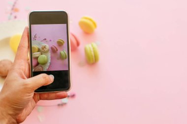El telefon tutarak ve lezzetli renkli macarons fotoğrafı trendy pastel pembe kağıt düz yatıyordu tabağa alarak. metin için yer. modern gıda fotoğrafçılığı kavramı. instagram Fotoğraf Atölyesi