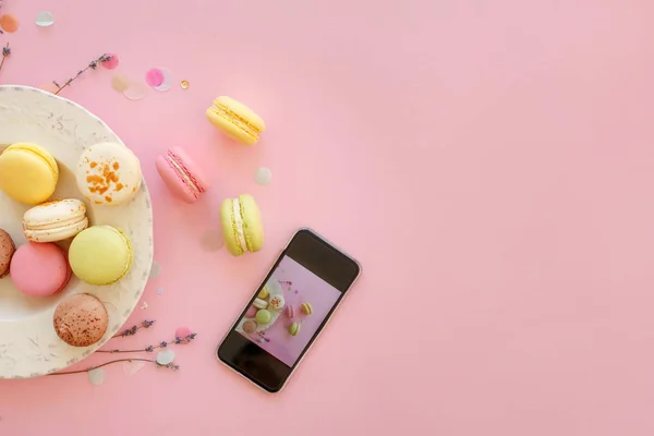 instagram food photography of tasty colorful macarons in plate and phone on trendy pastel pink paper flat lay. space for text. modern photo workshop. holiday catering